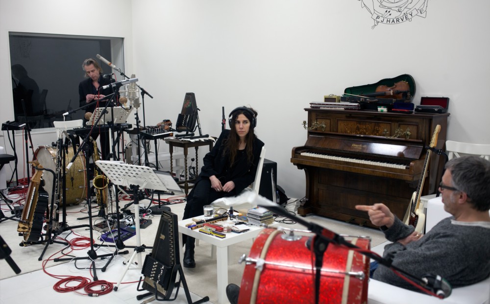 Musician PJ Harvey sits in a white recording studio with two men; there are instruments and microphones all over.