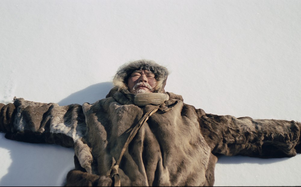 An Arctic indigenous man in a fur coat lays on snow.