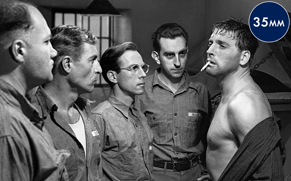 A group of men stand around Burt Lancaster, who is smoking a cigarette and has his shirt unbuttoned.