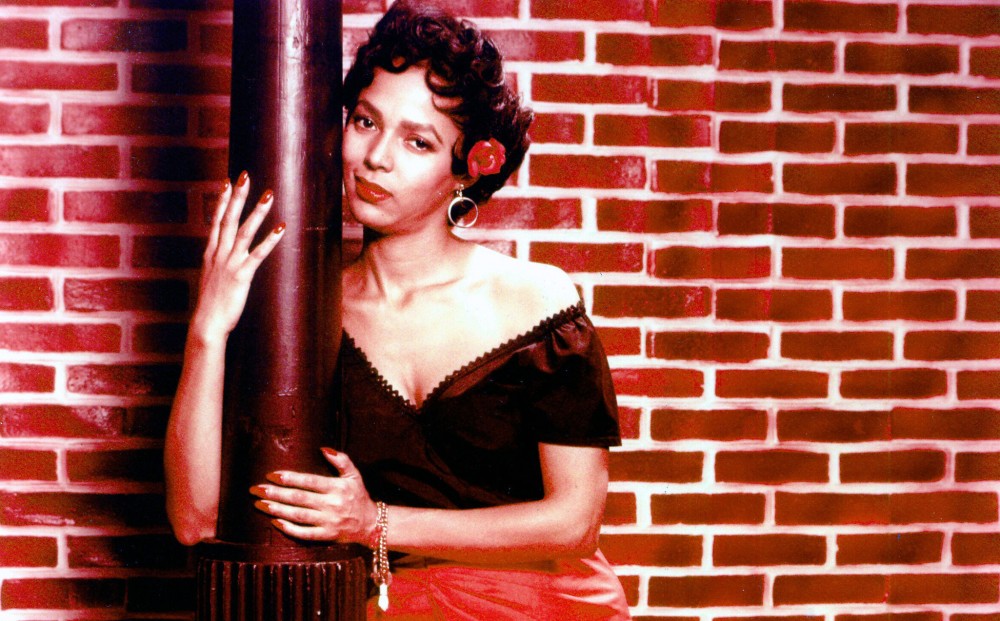Actor Dorothy Dandridge leans on and wraps her arms around a pole, against the background of a red brick wall.