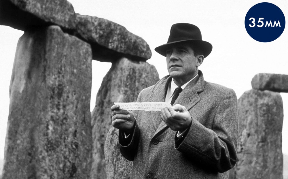 Actor Dana Andrews holds up a strip of paper that appears to be a clue, with Stongehenge in the background.