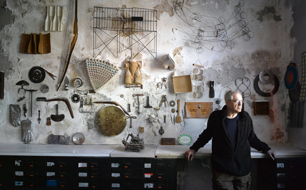 Artist and subject Jay Maisel leans against filing cabinets in his studio; objects like tools, gloves, and wires are mounted on the wall behind him.