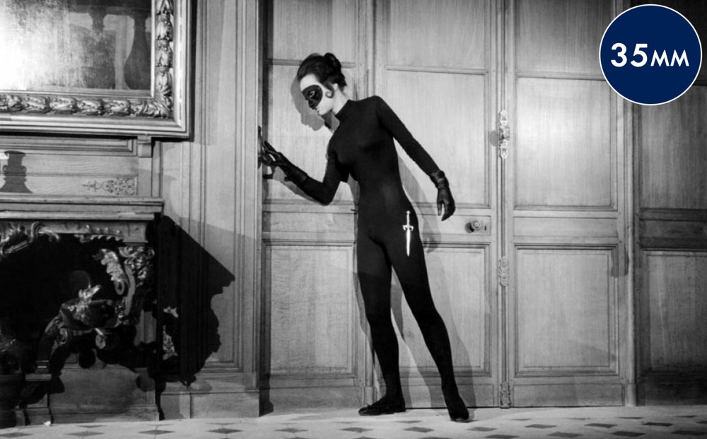 Actor  Francine Bergé lingers by a doorway, incognito in a black catsuit and black eye mask, with a dagger at her hip.