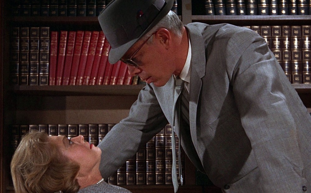 Actor Lee Marvin, wearing sunglasses, a hat, a suit, and tie, hovers menacingly over a frightened-looking woman.