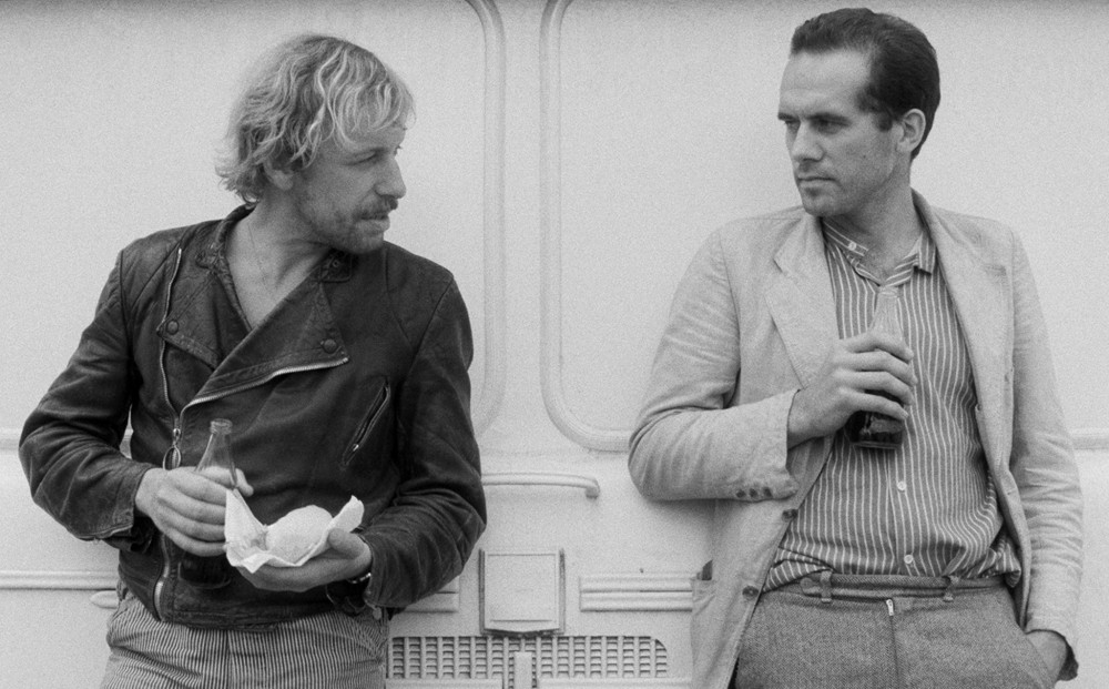 Actors Rüdiger Vogler and Hanns Zischler converse, each holding a beverage.