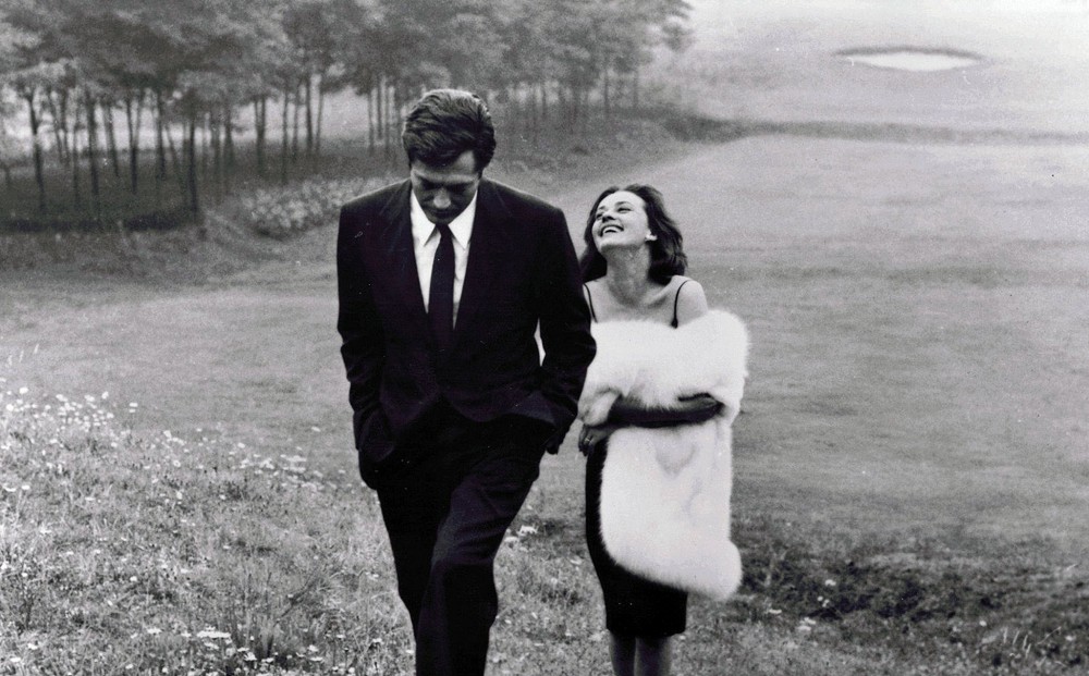 Actors Marcello Mastroianni and Jeanne Moreau walk in an abandoned golf course, both dressed in fancy party clothes.