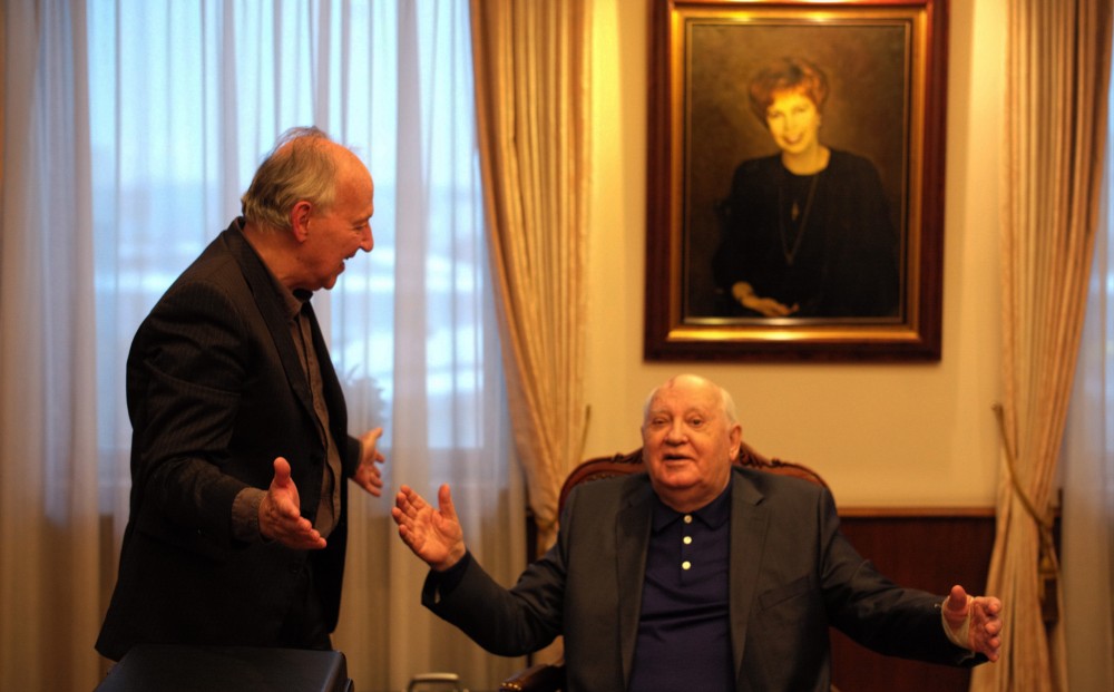 Mikhail Gorbachev, seated, and Werner Herzog, standing, gesture towards each other with their arms spread.