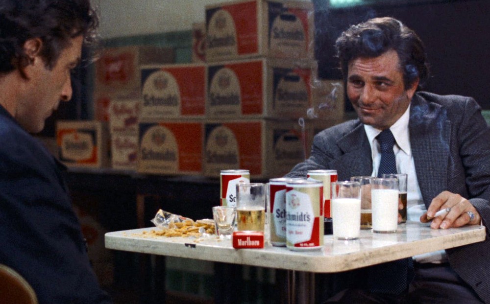 Actors  John Cassavetes and Peter Falk sit at a table that is covered in cans and cups of beer, and cups of milk; the latter smokes a cigarette.