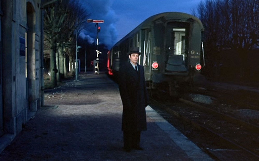 Actor Alain Delon waits on the platform at a train station at twilight.