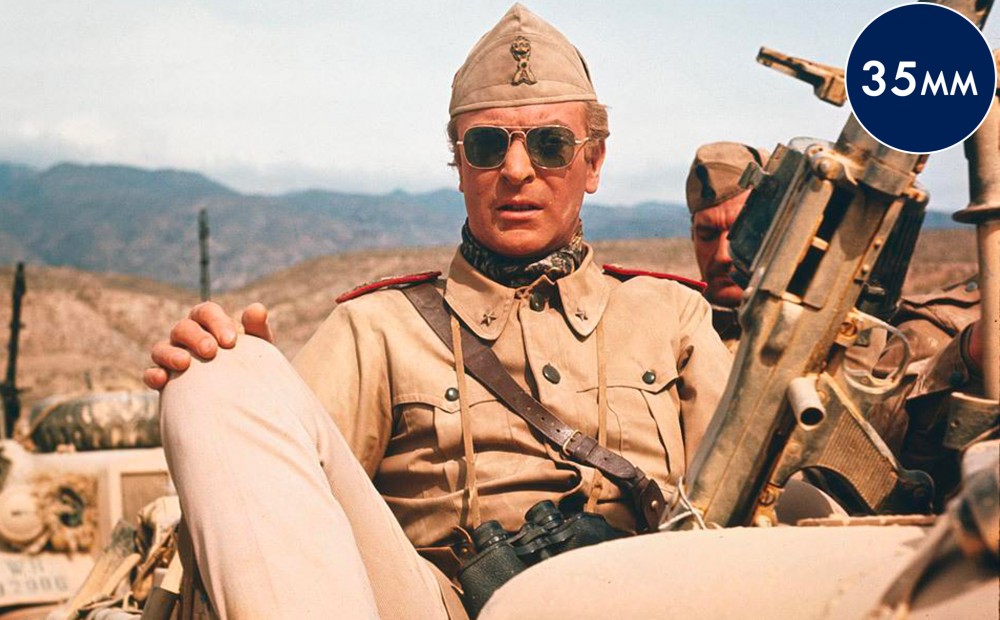 Actor Michael Caine sits on a military vehicle with a desert landscape.