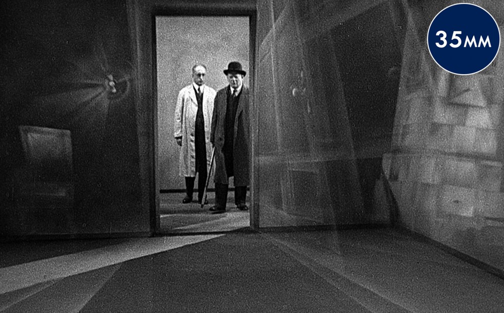 Two men stand in a doorway, one in a white coat and one in a black coat and bowler hat.