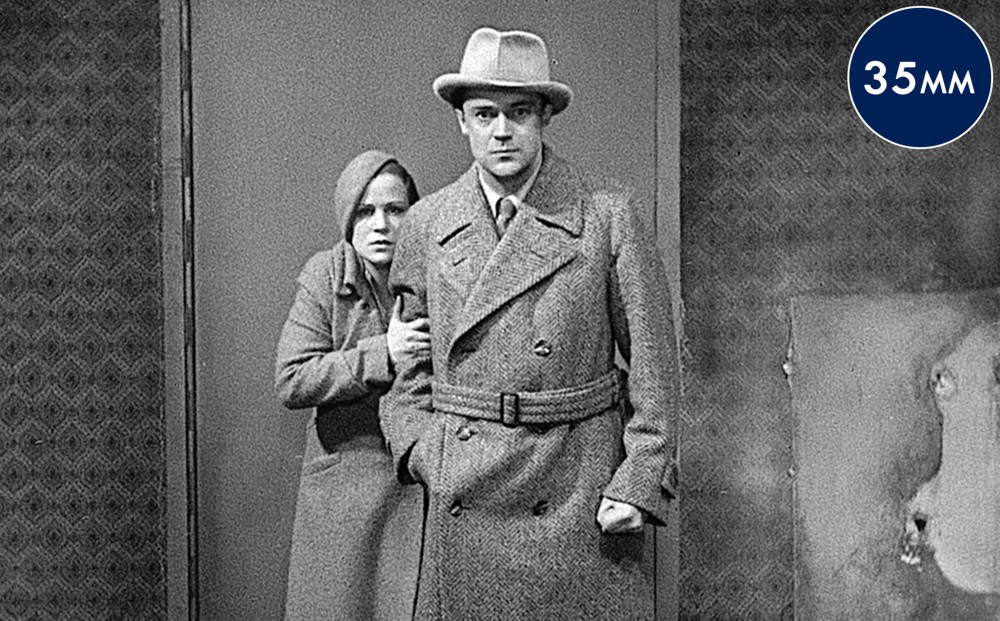 A man and woman stand in front of a door. He has one hand in his pocket and the other is clenched into a fist; she clutches his arm, stands slightly behind him, and looks frightened.