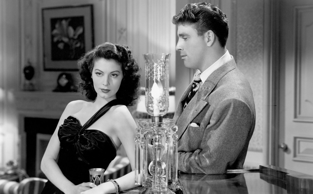 Actor Ava Gardner looks off into the distance in a living room; Burt Lancaster looks at her.