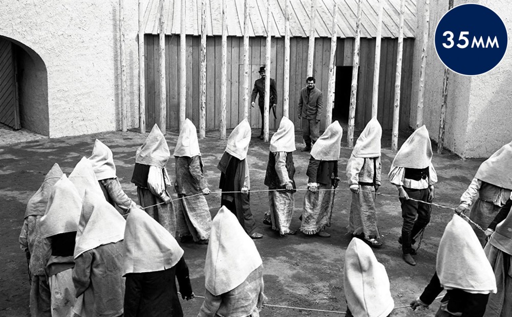 A group of prisoners with hoods over their heads all hold on to a rope and walk in a circle.