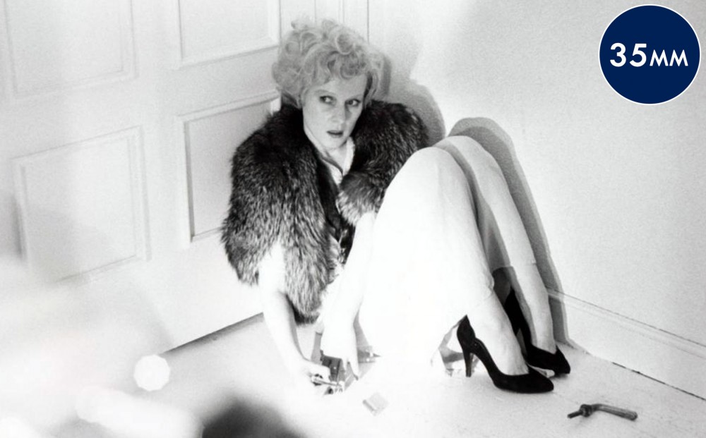 Actor Rosel Zech, wearing a fur stole and heels, sits on the ground, crouched against a doorway.