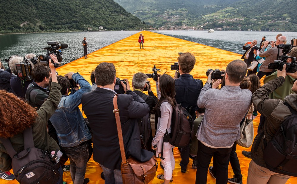 A crowd of photographers snap photographs of Christo, who stands at a distance, all on 'The Floating Piers.'