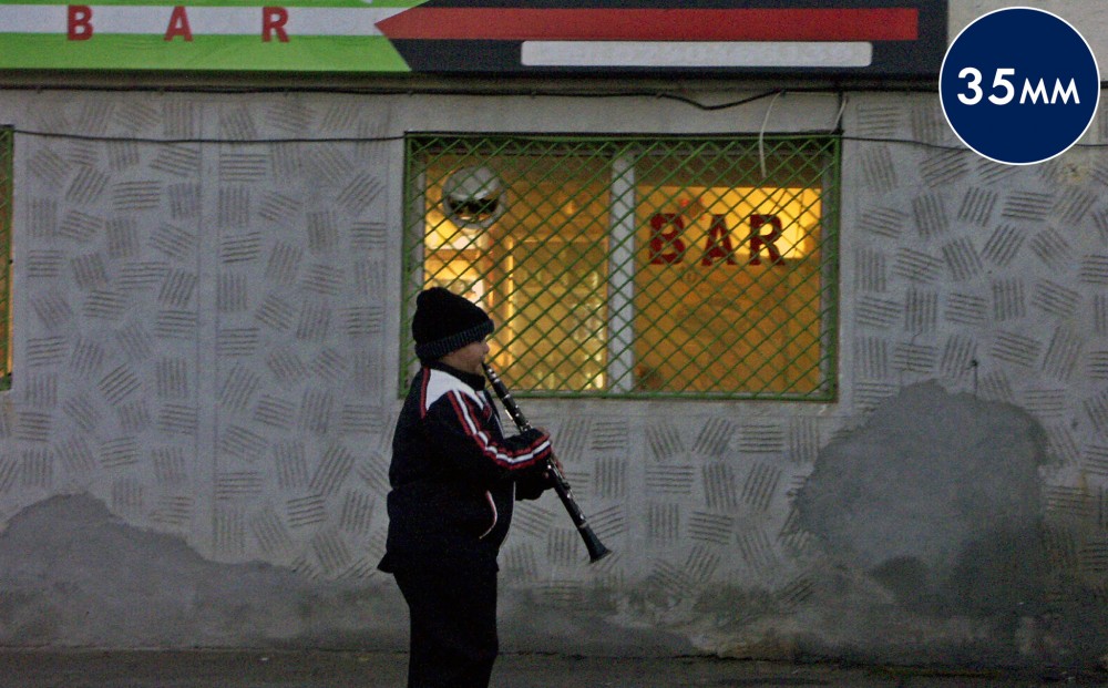 A young boy in a jacket and hat plays the clarinet outside of a bar.