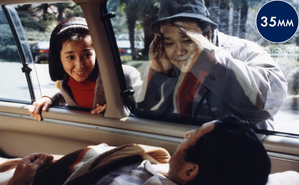 Two people look into a car through its window; a man appears to be sleeping inside.