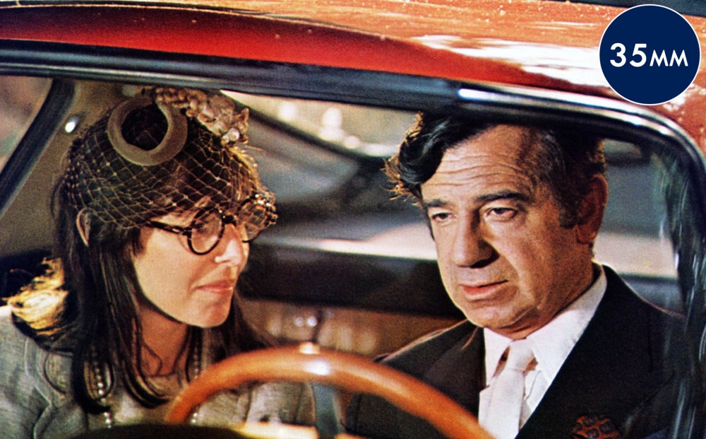 Actors Walter Matthau and Elaine May sit and talk in a car, with him at the wheel.