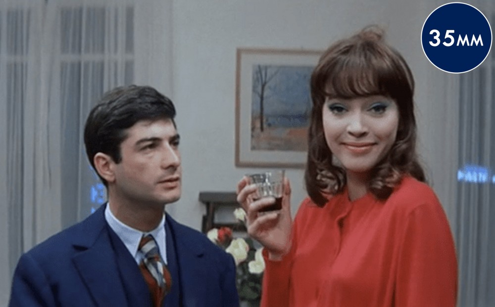 Actor Anna Karina holds a glass and looks towards the camera; a man looks at her.