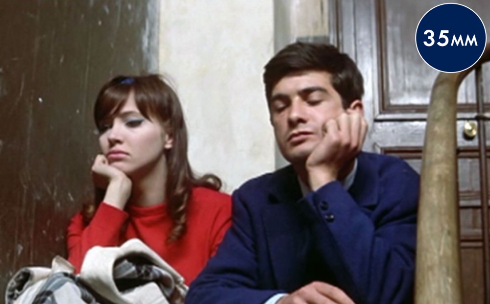 Actor Anna Karina and a man sit next to each other with their chins rested on their hands.