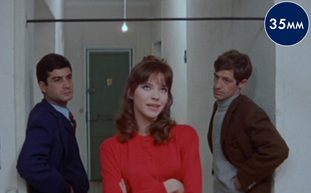 Two men look at actor Anna Karina, who has her arms crossed over her chest; all stand in a hallway.