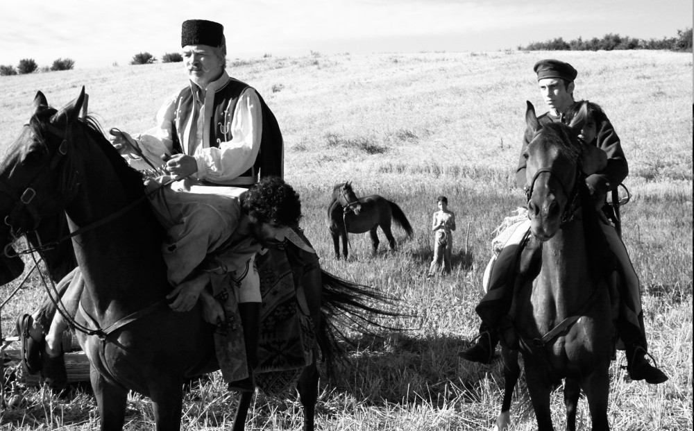 Black and white image of an older man on a horse next to a younger man on a horse; a shirtless child stands next to a horse in the vast field behind them.