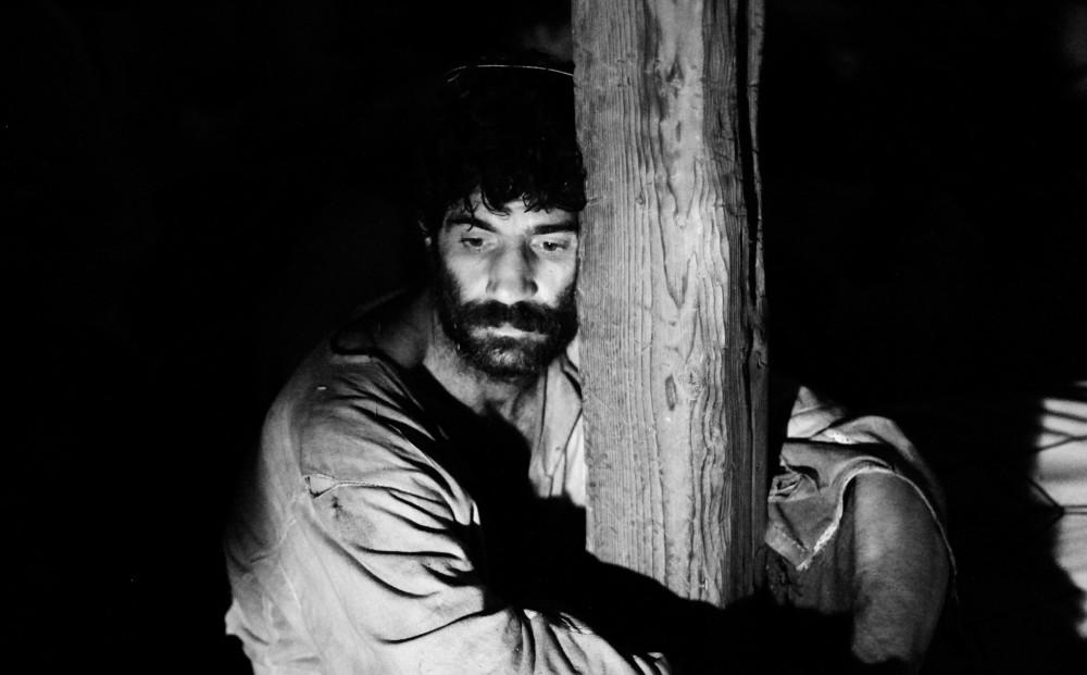 Black and white image of a man leaning against a wooden pole in the dark.