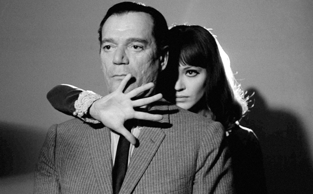 Actor Anna Karina stands behind a man, her arm over his right shoulder and her palm splayed out in front of his chin.
