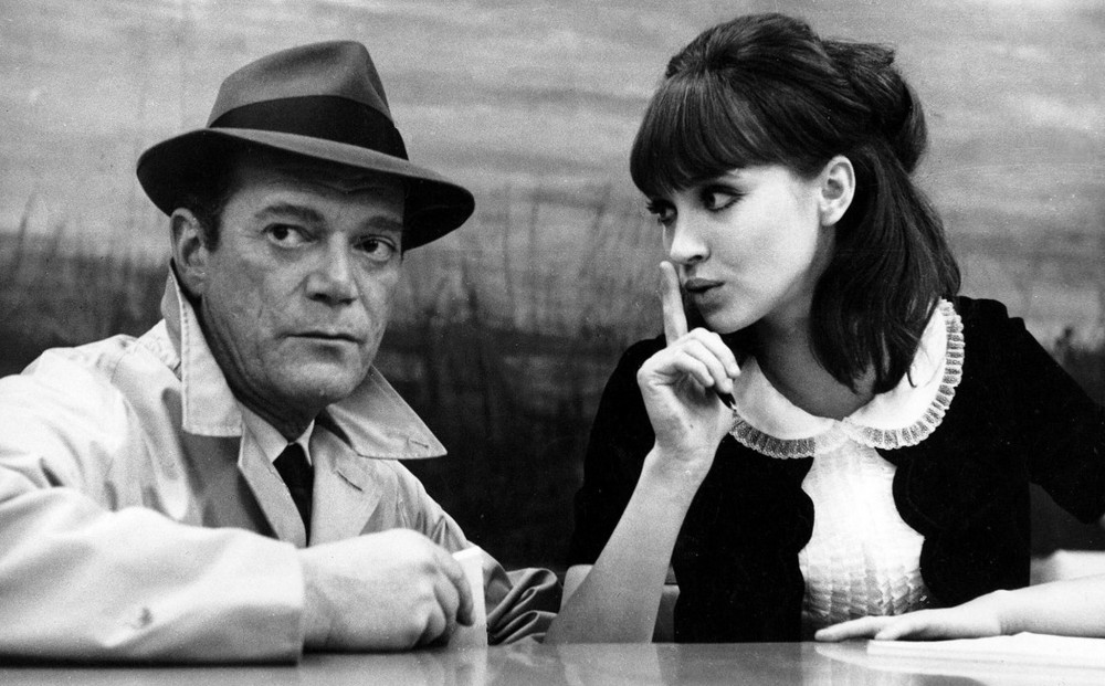 Actor Anna Karina sits at a table with a man, and holds her index finger over her mouth in a 