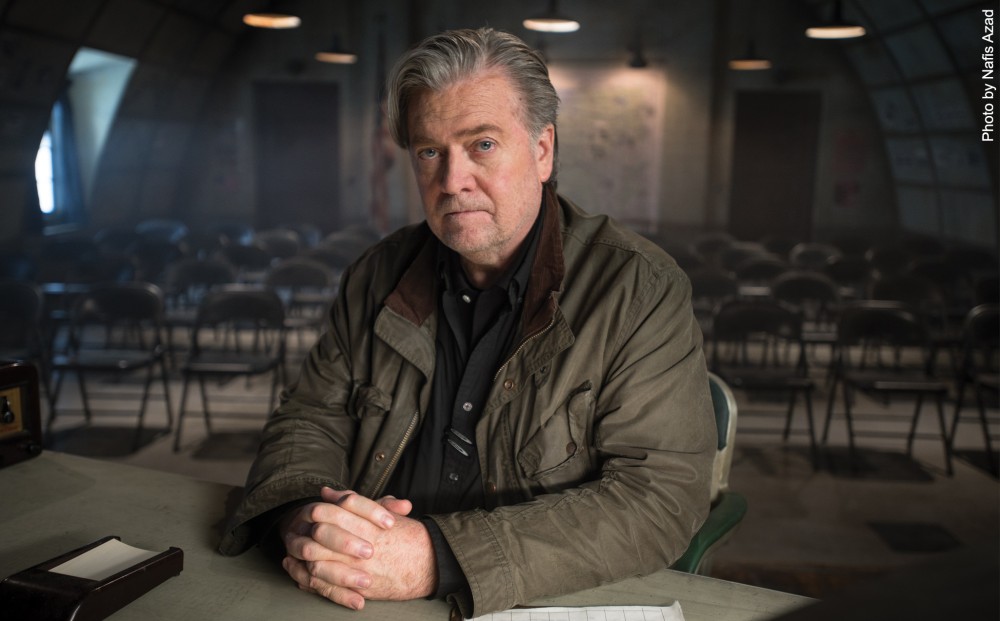 Steve Bannon sits at a desk in a darkened room, with rows of folding chairs behind him.
