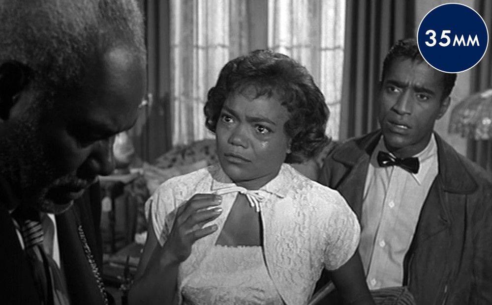 Actor Eartha Kitt, with tears in her eyes, and a man who looks upset, both gaze at an older man, in profile with downcast eyes.