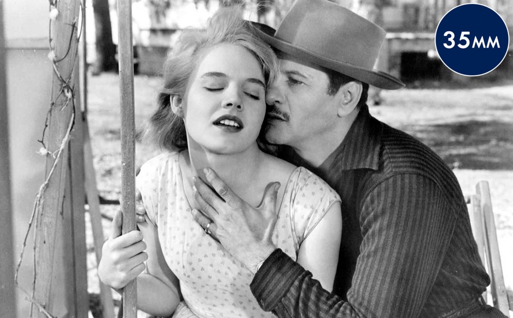 Actors Eli Wallach and Carroll Baker sit next to each other on a bench or swing; his hand is over her chest as he appears to whisper in her ear.