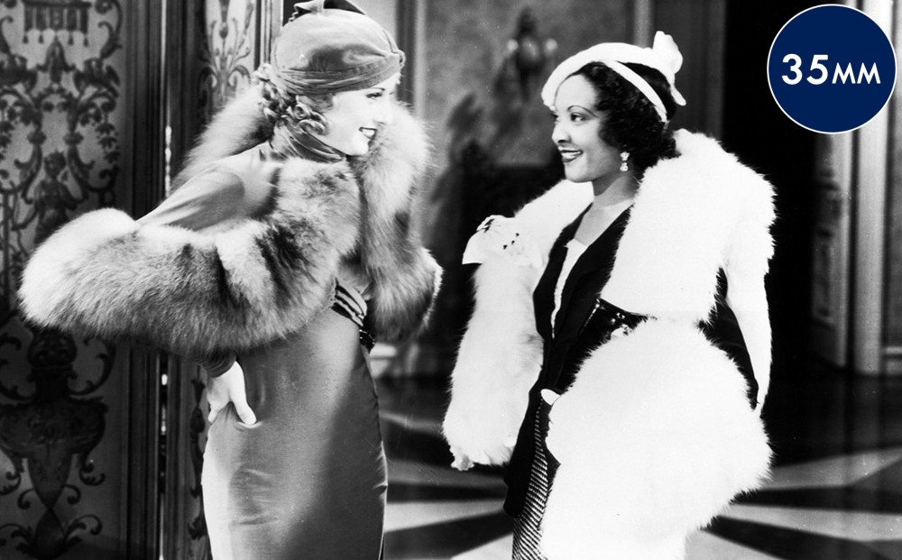 Actors Barbara Stanwyck and Theresa Harris stand and smile at each other, both dressed in fine clothes with fur stoles.