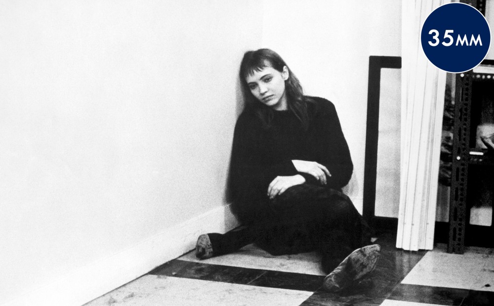 Anna Karina sits on the floor, leaning against a wall and looking sad.