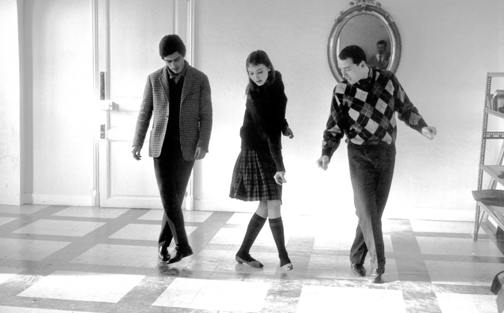 Actor Anna Karina dances with two men, one on either side of her.