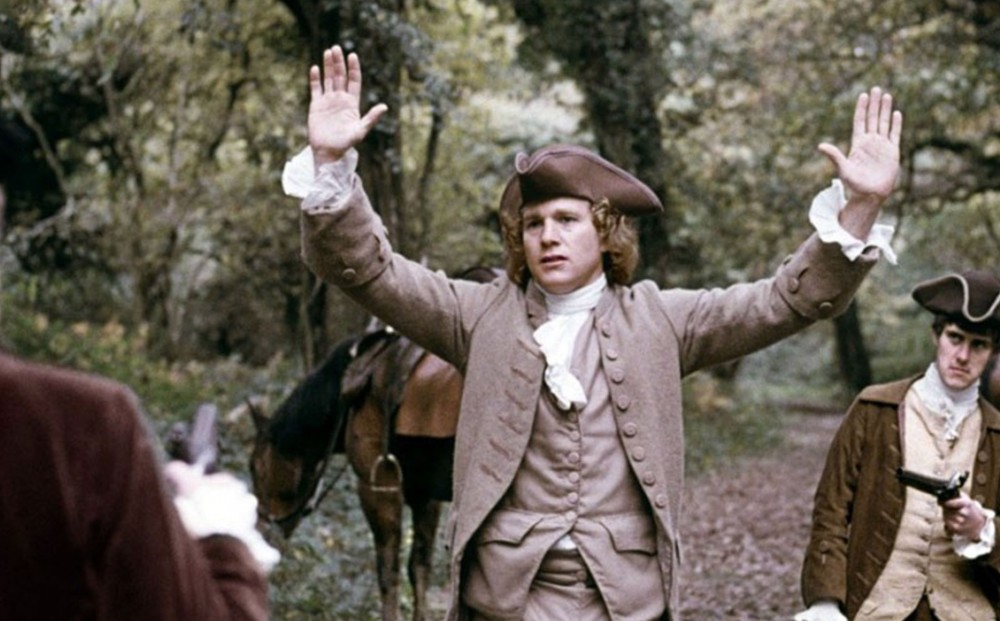 Actor Ryan O'Neal stands in a forest with his hands up; two men point their guns at him.