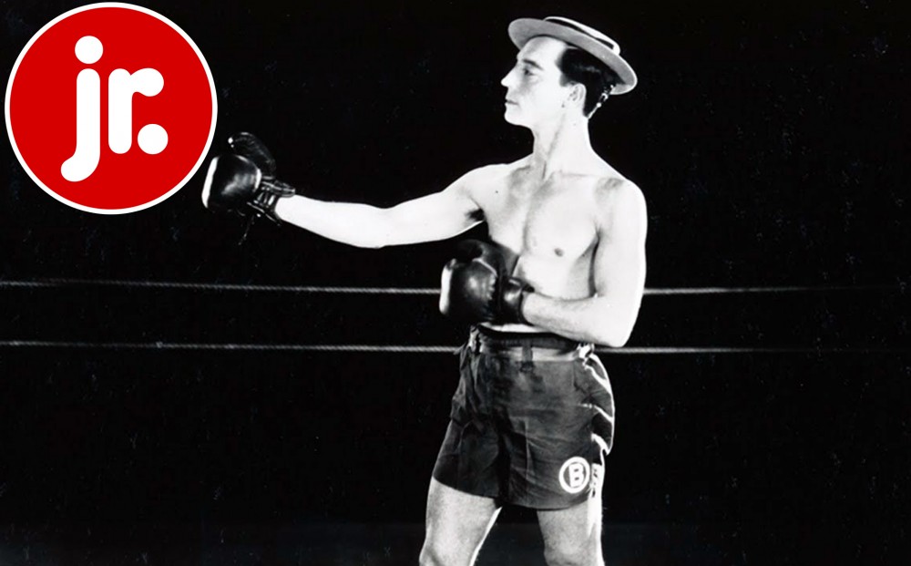 Buster Keaton stands in a boxing ring wearing boxing shorts and boxing gloves.
