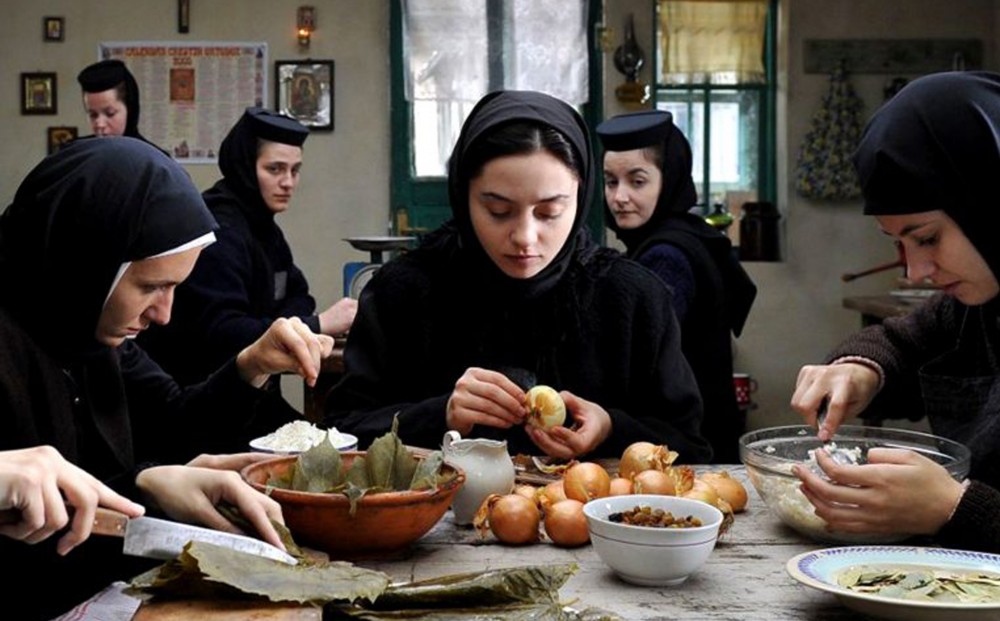 Nuns in a kitchen sit at a table, chopping leaves, peeling onions, and doing other food preparation tasks.