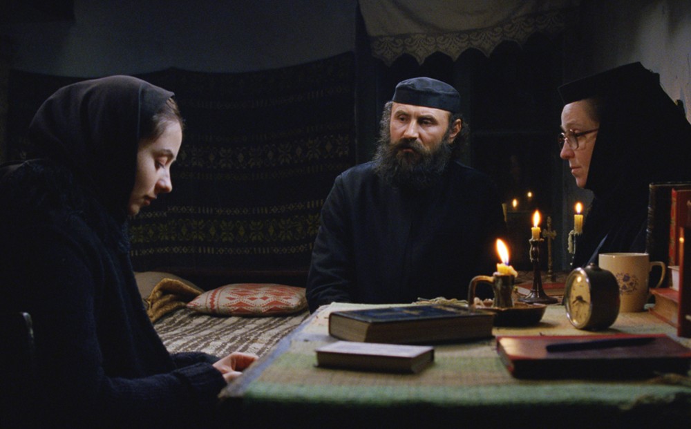 A nun and a priest sit with a young woman in a room with books and candles on a table.