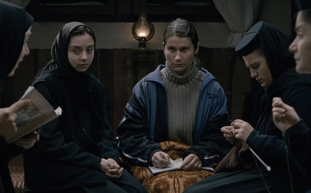 Two young women sit on their bed, two other nuns are reading and knitting next to them.