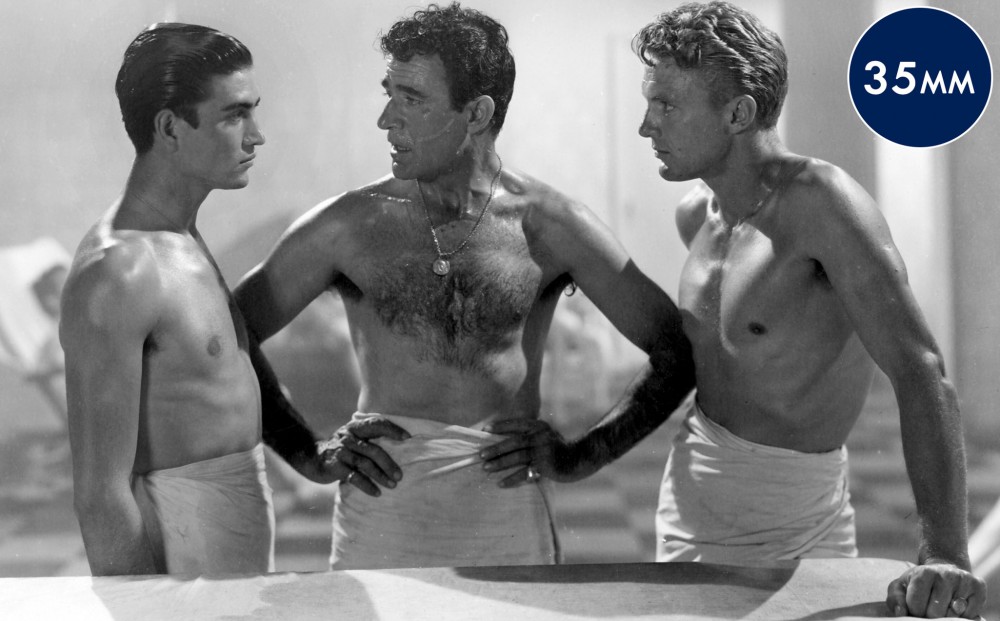 Three men wearing only towels around their waists are engaged in a serious discussion.