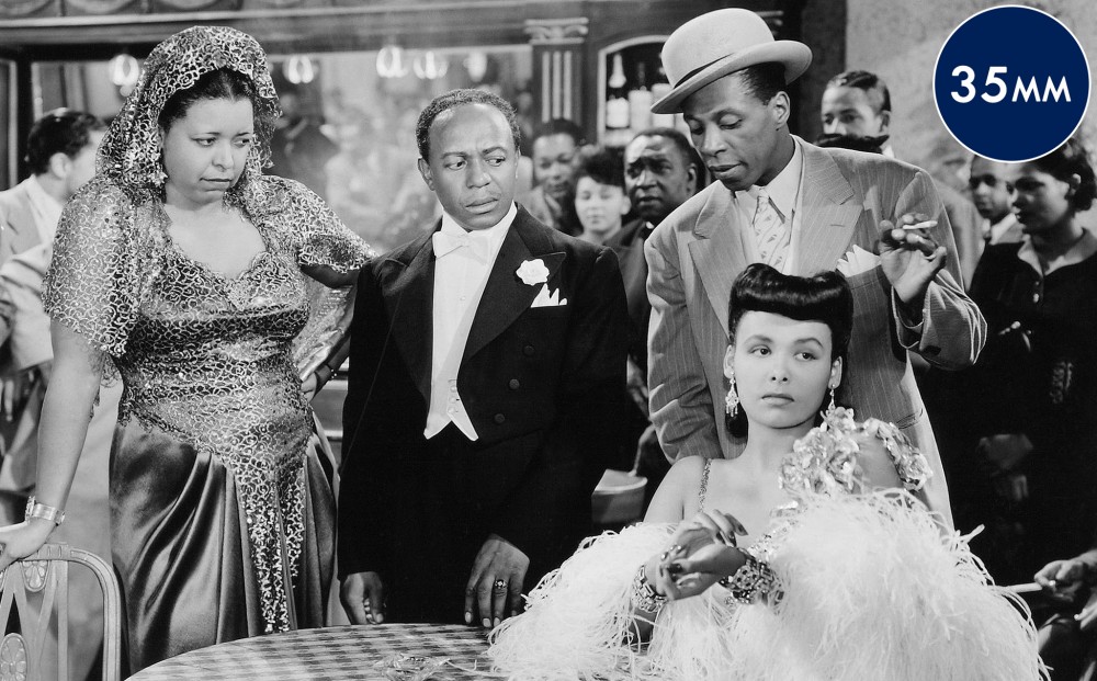 Actor Lena Horne sits at a table in a nightclub, while another woman and two men stand beside it, looking at her with consternation. All are dressed in fine evening wear.