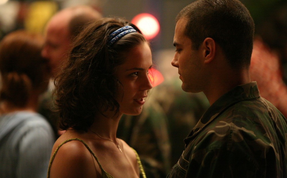 A man wearing a green camouflage shirt and a woman stand close to and face each other, on what looks to be a dance floor.