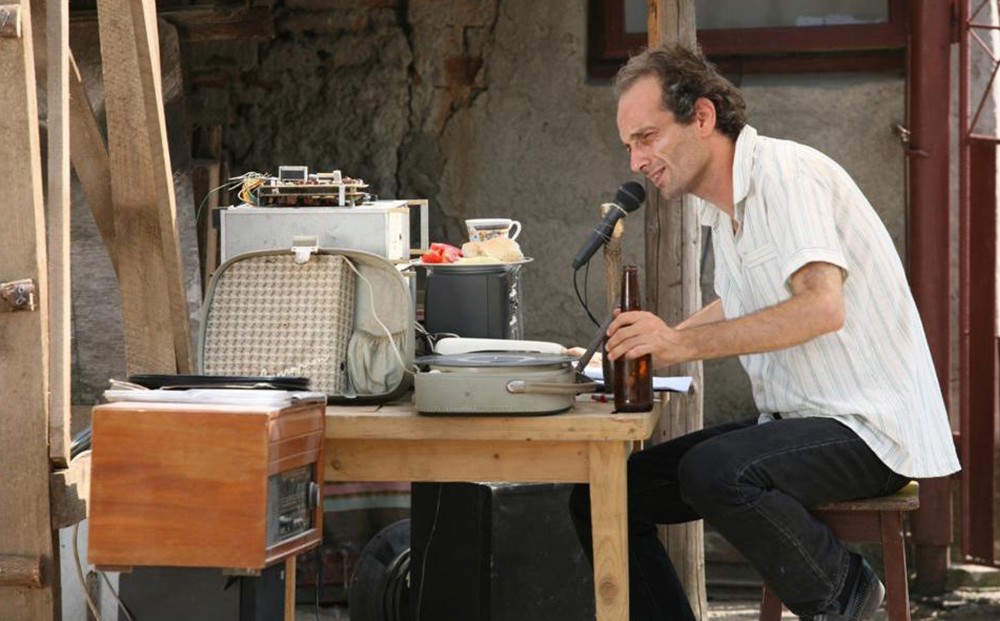 A man seated at a wood table covered in junk holds a beer bottle in one hand, and speaks into a microphone that is being propped up on a stick, to which it is affixed by twine or a rubber band.