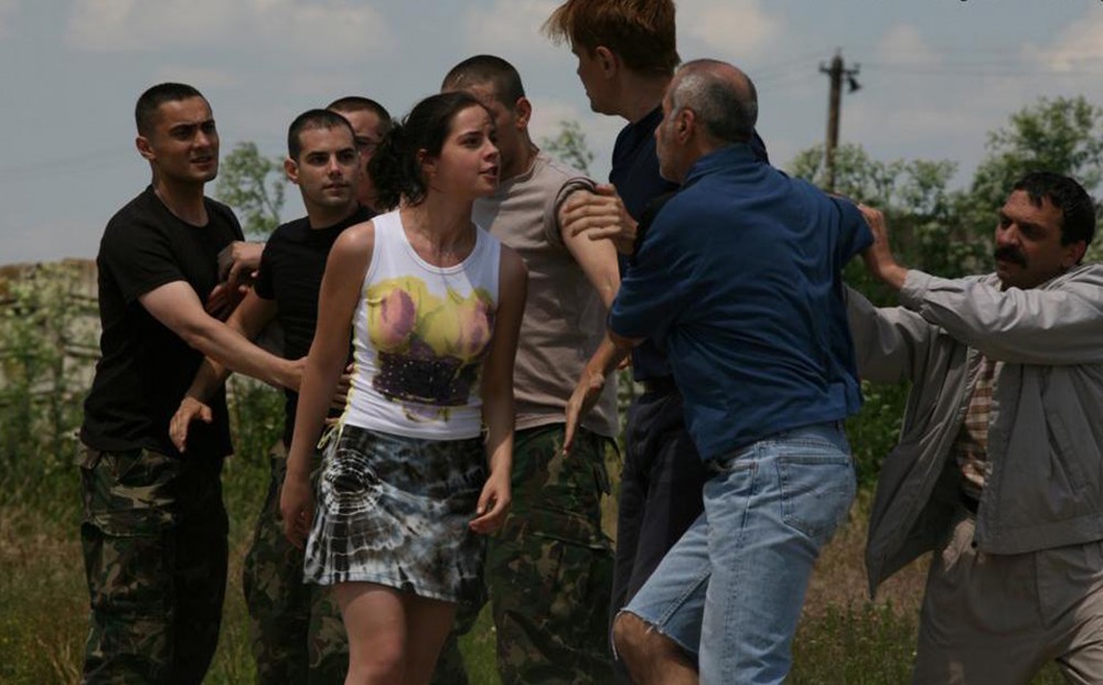 A group of eight people, including four men in green camouflage pants, are engaged in an altercation, with some trying to hold others back from fighting.