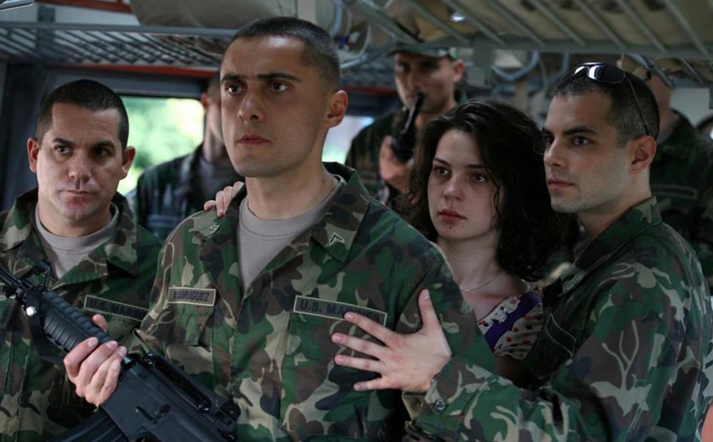 A man in U.S. marine green army fatigues holds a gun, flanked by other marines and a woman just behind him.