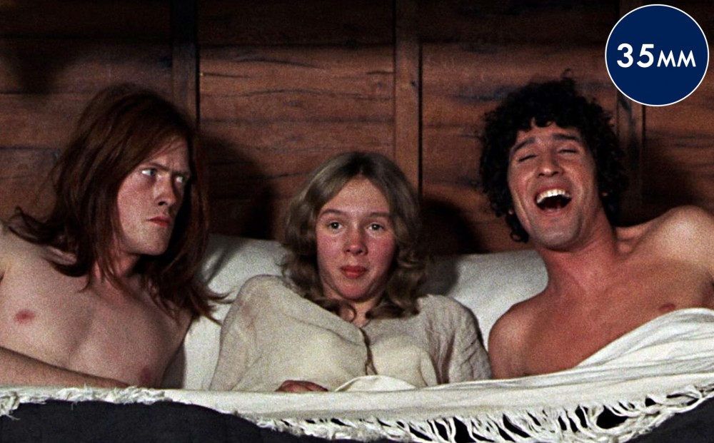 A women wearing a nightgown lays on a bed between two shirtless men—one looks angry at the other, who is laughing.