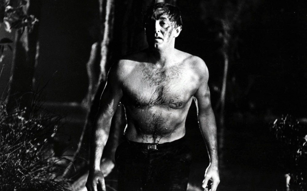 A shirtless, wet, and dirty Robert Mitchum walks towards the camera.