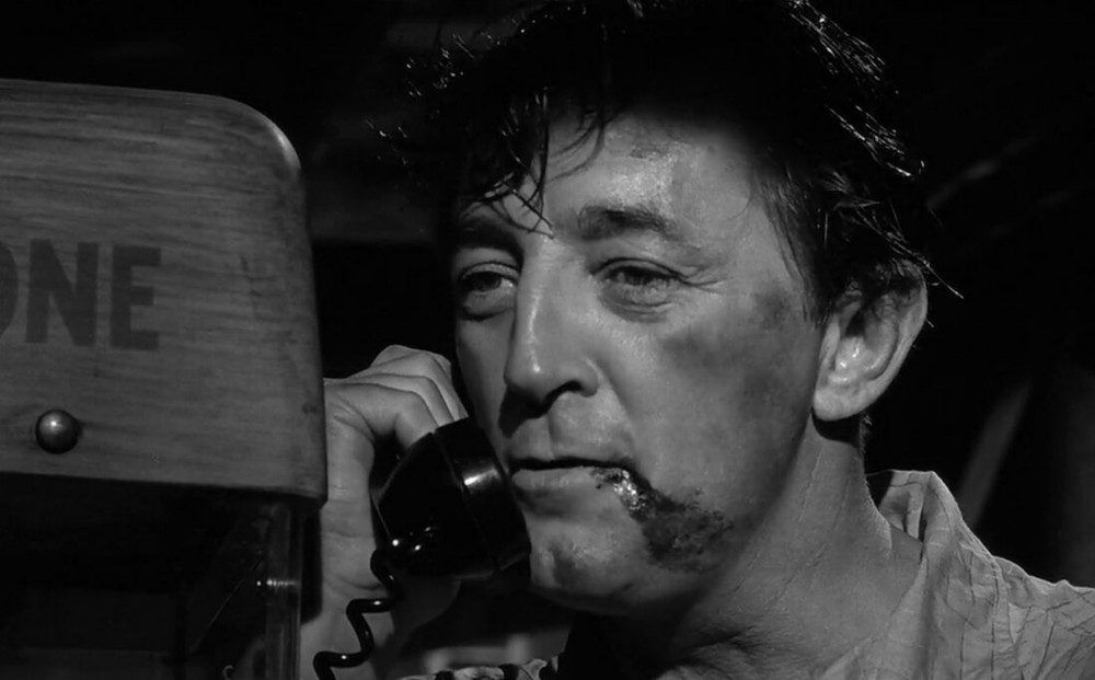 Actor Robert Mitchum speaks into a pay phone; he has a large, bloody scrape near his mouth.
